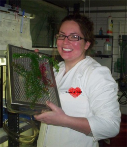 Emily, wearing white lab coat and holding up green fern-like frond on screen.